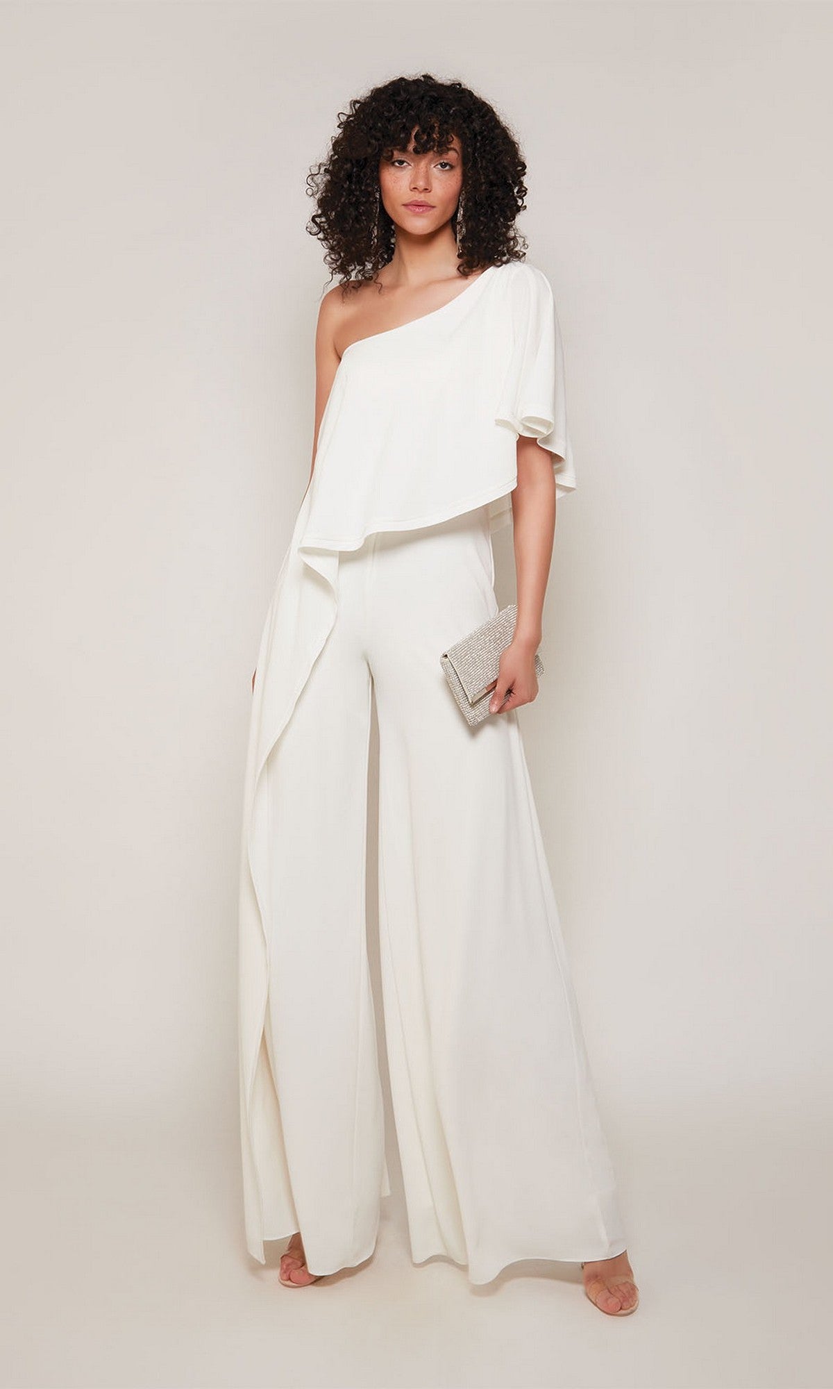 A woman wears the Alyce 70049 Long Formal Jumpsuit with an off-shoulder wide-leg design, holding a small silver clutch and standing in front of a plain light background.